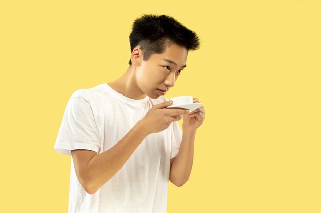 Retrato de medio cuerpo del joven coreano sobre fondo amarillo de estudio. Modelo masculino en camisa blanca. Beber café, sentirse feliz. Concepto de emociones humanas, expresión facial. Vista frontal. Colores de moda.