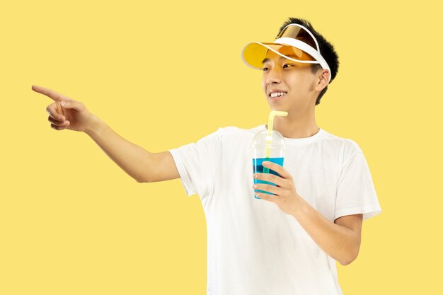 Retrato de medio cuerpo del joven coreano sobre fondo amarillo de estudio. Modelo masculino con camisa blanca y gorra amarilla. Beber cóctel. Concepto de emociones humanas, expresión, verano, vacaciones, fin de semana.