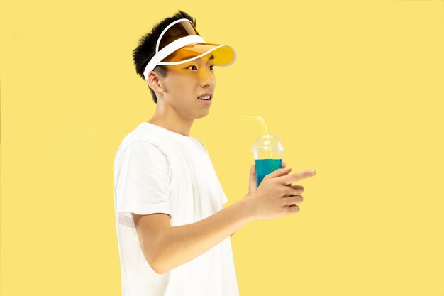 Retrato de medio cuerpo del joven coreano sobre fondo amarillo de estudio. Modelo masculino con camisa blanca y gorra amarilla. Beber cóctel. Concepto de emociones humanas, expresión, verano, vacaciones, fin de semana.