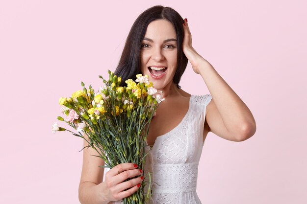 Retrato de medio cuerpo de mujer bonita morena con ramo de flores, riendo mientras mira directamente a la cámara, mantiene la mano sobre la cabeza
