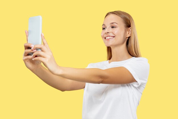 Retrato de medio cuerpo de la mujer joven caucásica sobre fondo amarillo de estudio. Modelo de mujer hermosa en camisa blanca. Concepto de emociones humanas, expresión facial. Sonriendo, haciendo contenido para selfies o vlogs.