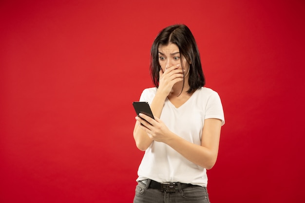 Retrato de medio cuerpo de la mujer joven caucásica sobre fondo rojo de estudio. Modelo de mujer hermosa en camisa blanca. Concepto de emociones humanas, expresión facial. Compra en línea, paga la factura, parece sorprendido.