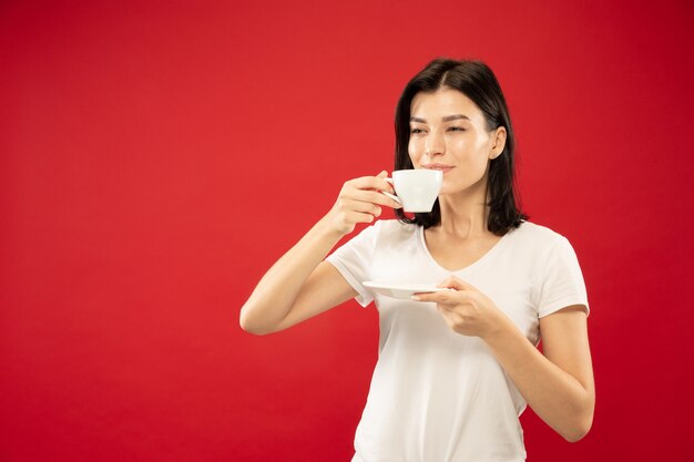 Retrato de medio cuerpo de la mujer joven caucásica sobre fondo rojo de estudio. Modelo de mujer hermosa en camisa blanca. Concepto de emociones humanas, expresión facial. Disfruta tomando café o té, parece tranquilo.