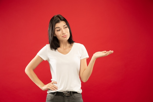 Retrato de medio cuerpo de la mujer joven caucásica sobre fondo rojo de estudio. Modelo de mujer hermosa en camisa blanca. Concepto de emociones humanas, expresión facial. Mostrando algo de incertidumbre, eligiendo.