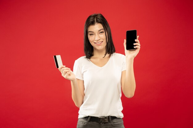 Retrato de medio cuerpo de la mujer joven caucásica sobre fondo rojo de estudio. Modelo de mujer hermosa en camisa blanca. Concepto de emociones humanas, expresión facial, ventas. Pago de facturas o compras online.