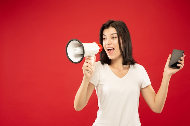 Retrato de medio cuerpo de la mujer joven caucásica sobre fondo rojo de estudio. Modelo de mujer hermosa en camisa. Concepto de emociones humanas, expresión facial, ventas. Sosteniendo el teléfono y llamando en boca en paz.