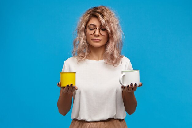 Retrato de moda adorable joven secretaria en anteojos redondos hoding dos tazas en sus manos