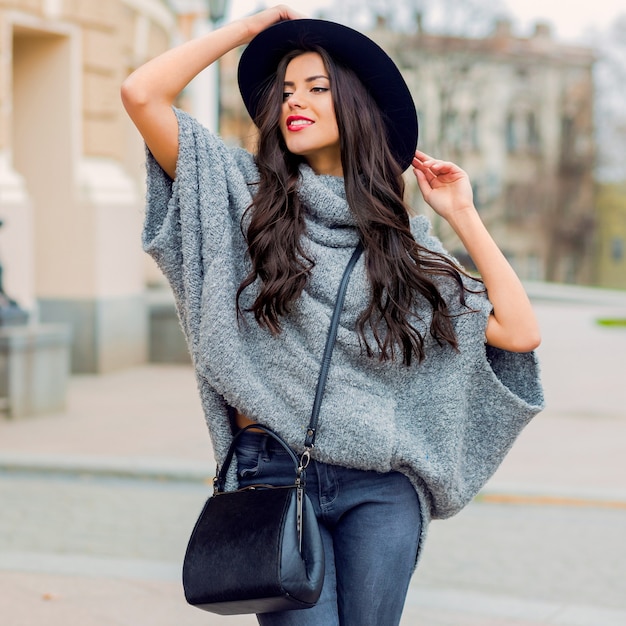 Retrato de moda al aire libre de glamour sensual elegante joven vistiendo ropa de otoño, sombrero negro, suéter gris y bolso de cuero. Labios rojos brillantes. Ciudad vieja de fondo.