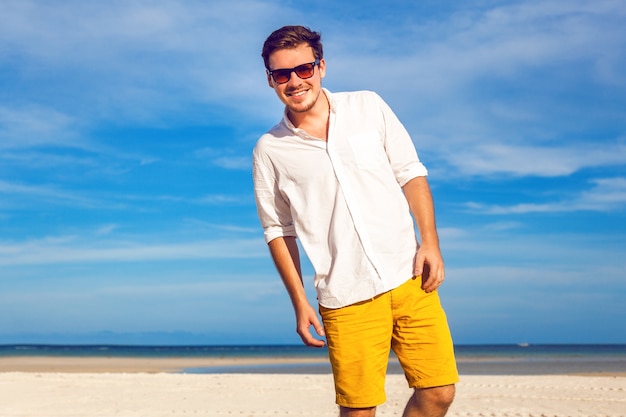 Retrato de moda al aire libre de hombre guapo posando en la increíble playa tropical, en un bonito día soleado, hermosa vista sobre el cielo azul y el océano, vestido con camisa blanca clásica y gafas de sol de color amarillo casual.