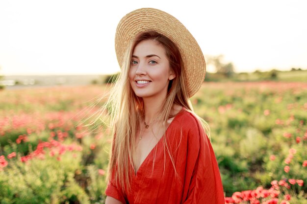 Retrato de moda al aire libre de impresionante mujer rubia posando mientras camina en el increíble campo de amapolas en una cálida noche de verano. Con sombrero de paja, bolso de moda y vestido rojo.