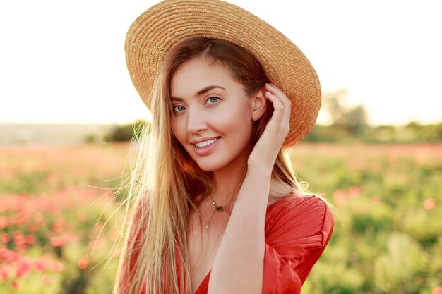 Retrato de moda al aire libre de impresionante mujer rubia posando mientras camina en el increíble campo de amapolas en una cálida noche de verano. Con sombrero de paja, bolso de moda y vestido rojo.