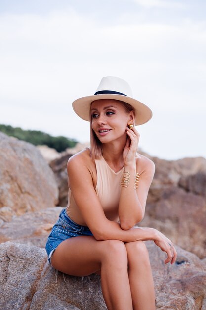 Retrato de moda al aire libre de una joven europea en mono beige, pantalones cortos de jean azul y sombrero con estilo clásico en la cálida luz del atardecer de la playa rocosa tropical