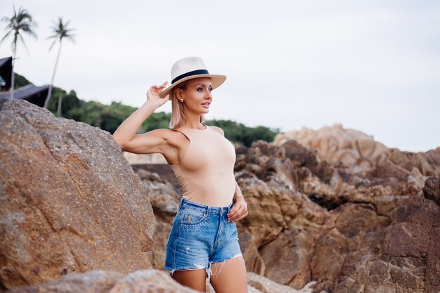 Retrato de moda al aire libre de una joven europea en mono beige, pantalones cortos de jean azul y sombrero con estilo clásico en la cálida luz del atardecer de la playa rocosa tropical