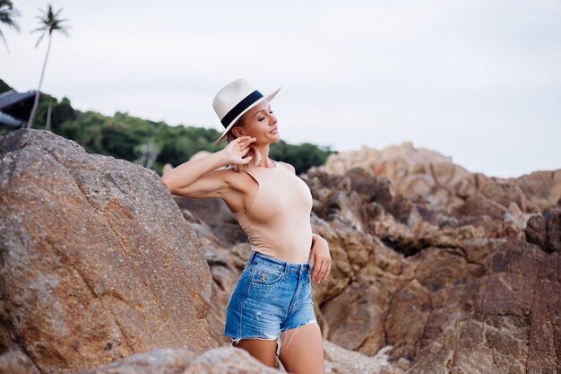 Retrato de moda al aire libre de una joven europea en mono beige, pantalones cortos de jean azul y sombrero con estilo clásico en la cálida luz del atardecer de la playa rocosa tropical