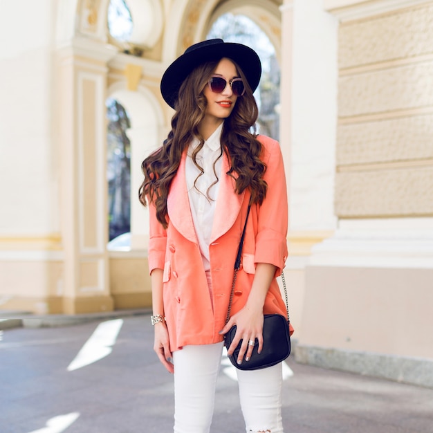 Retrato de moda al aire libre de moda de mujer casual con sombrero negro, traje rosa, blusa blanca posando en la calle vieja
