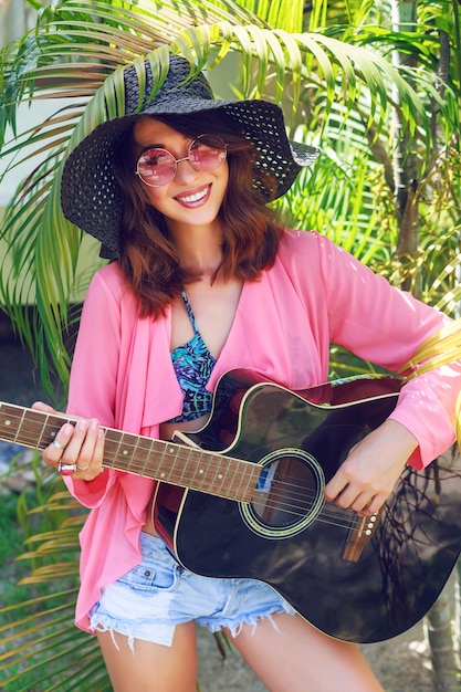 Retrato de moda al aire libre de mujer hippie muy sonriente feliz sentado en el césped y sosteniendo la guitarra acústica. País tropical caliente, fondo verde. Traje de verano con sombrero y gafas de sol rosas.