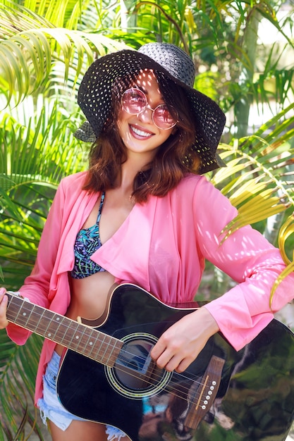 Retrato de moda al aire libre de mujer hippie muy sonriente feliz sentado en el césped y sosteniendo la guitarra acústica. País tropical caliente, fondo verde. Traje de verano con sombrero y gafas de sol rosas.