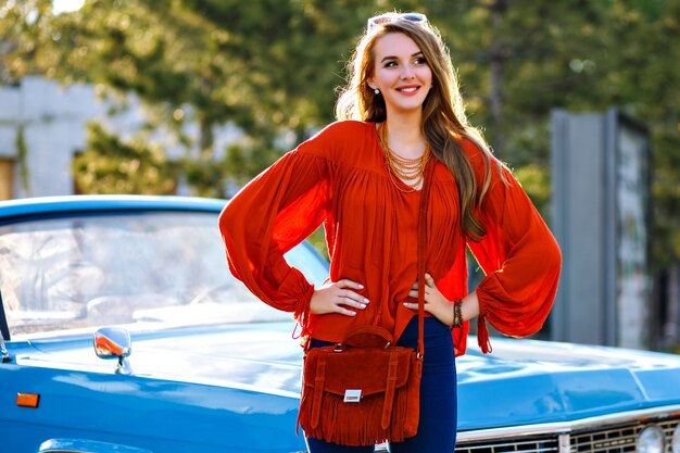 Retrato de moda al aire libre de una mujer joven elegante con increíbles pelos largos rubios y una cara bonita sonriendo y disfrutando de un día soleado, posando junto a un coche de época azul, un traje moderno de glamour boho, mal y joyas.