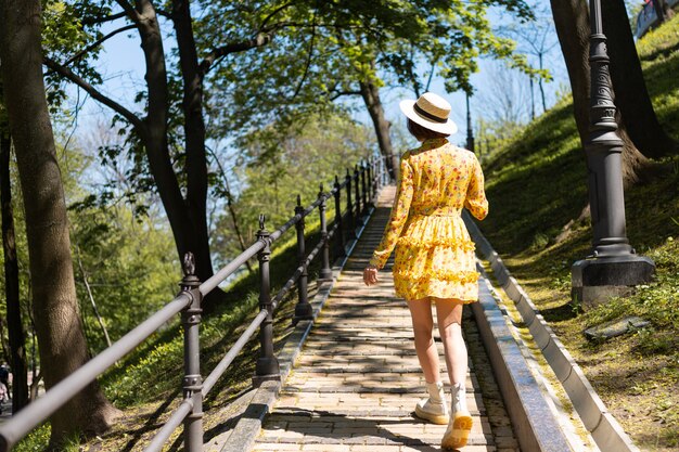 Retrato de moda al aire libre de mujer en vestido amarillo de verano y sombrero caminando en el camino en la vista posterior del parque