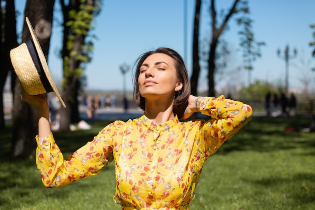 Foto gratuita retrato de moda al aire libre de mujer en vestido amarillo de verano y sombrero sentado en la hierba en el parque