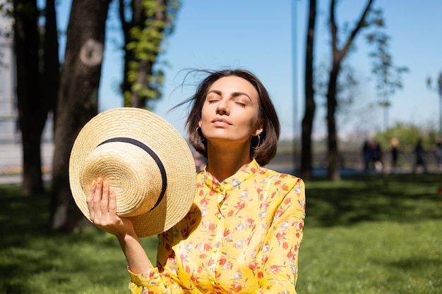 Retrato de moda al aire libre de mujer en vestido amarillo de verano y sombrero sentado en la hierba en el parque