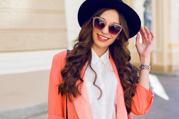 Retrato de moda de altura al aire libre de mujer casual elegante sexy con sombrero negro, traje rosa, blusa blanca posando en la calle vieja. Primavera, otoño día soleado. Peinado ondulado