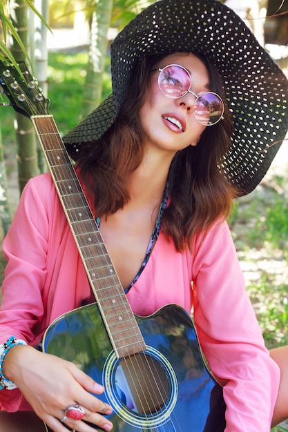 Retrato de moda de chica hermosa con maquillaje natural y pelos morenos mullidos posando en el jardín con guitarra. con sombrero y gafas de sol rosas redondas de moda.