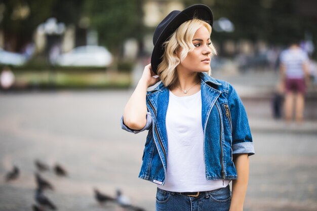 Retrato de moda chica rubia en el centro de la ciudad vestida con jeans suite y camiseta blanca y sombrero negro