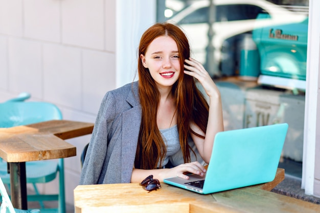 Retrato de moda de la ciudad al aire libre de la joven empresaria que trabaja en el café en la terraza en un día soleado, ropa casual elegante, detalles de menta, usando su computadora portátil, café, concepto de negocio