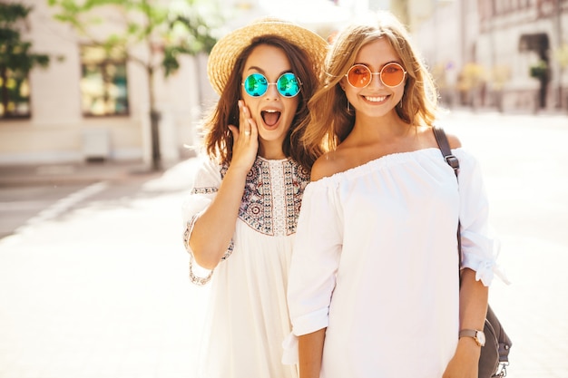 Retrato de moda de dos jóvenes hippie con estilo morena y rubia mujeres modelos. Mejores amigas en vestido blanco hipster de verano posando