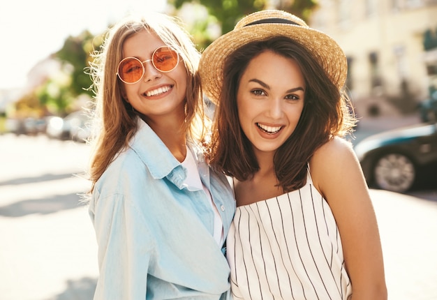 Retrato de moda de dos jóvenes sonrientes hippie con estilo morena y rubia mujeres modelos en día soleado de verano en ropa hipster posando en el fondo de la calle
