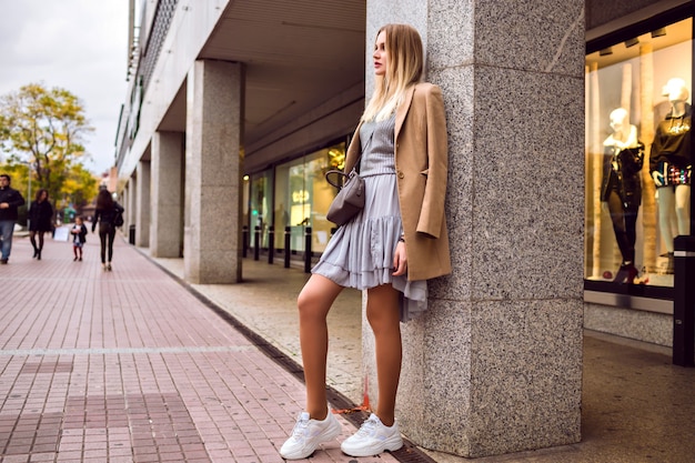 Retrato de moda de estilo callejero de mujer rubia elegante glamour posando en la calle europea cerca del centro comercial, elegante vestido suéter y abrigo de cachemira, colores tonificados, tiempo de primavera.