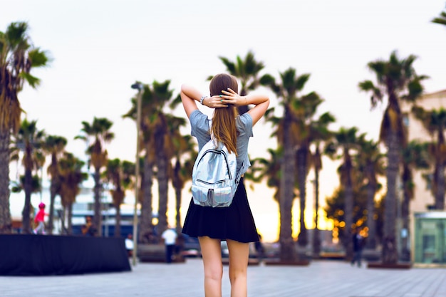 Retrato de moda de estilo de vida al aire libre de mujer joven inconformista caminando en Barcelona, viaje con mochila, elegante atuendo casual, atardecer, palmas, estudiante, peinado rubio, tiempo feliz, colores tonificados.