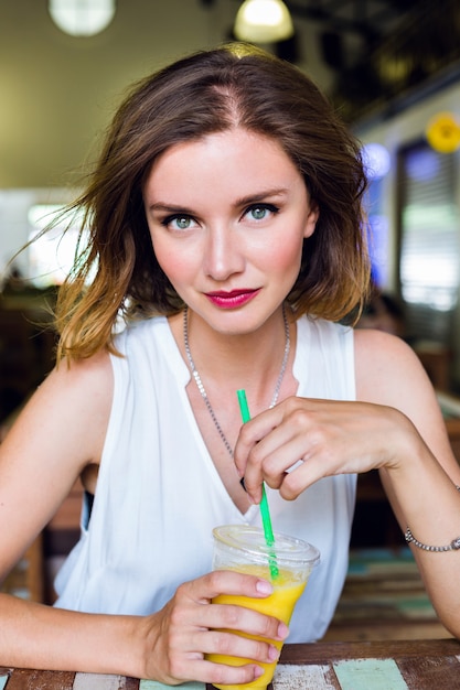 Retrato de moda de estilo de vida interior de mujer hermosa posando en el café, bebiendo jugo de mango sabroso saludable fresco, sonriendo, pasar un buen rato, maquillaje sexy brillante.