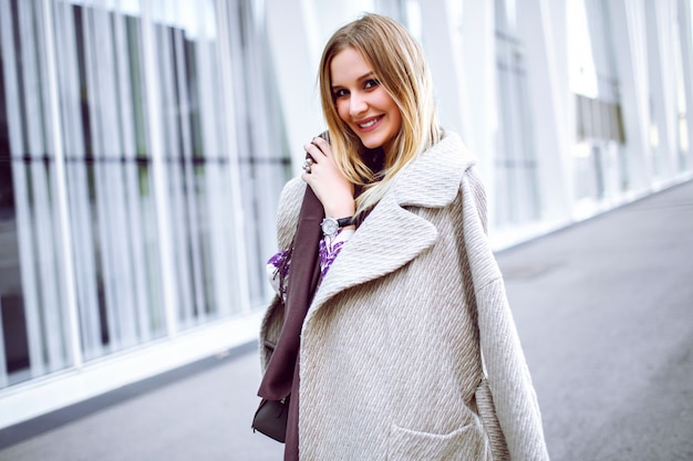 Retrato de moda de estilo de vida de una mujer muy elegante con un atuendo elegante y moderno, bufanda larga violeta, abrigo de lujo de cachemira y vestido midi, sonriendo y disfrutando, quédese frente al moderno centro de negocios