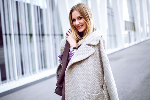 Retrato de moda de estilo de vida de una mujer muy elegante con un atuendo elegante y moderno, bufanda larga violeta, abrigo de lujo de cachemira y vestido midi, sonriendo y disfrutando, quédese frente al moderno centro de negocios