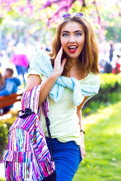 Retrato de moda de estilo de vida de primavera de linda mujer rubia posando en el parque de la ciudad, ropa casual brillante y mochila, emociones lindas, colores pastel soleados, vacaciones, al aire libre.