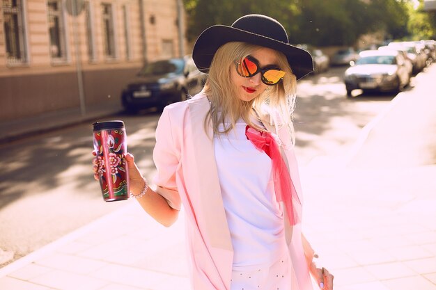 Retrato de moda de estilo de vida soleado de verano de joven elegante hipster mujer caminando en la calle, vistiendo lindo traje de moda, bebiendo café con leche caliente