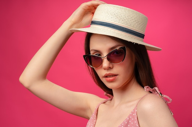 Retrato de moda de estudio de una mujer joven y atractiva con sombrero y gafas contra el fondo rosa