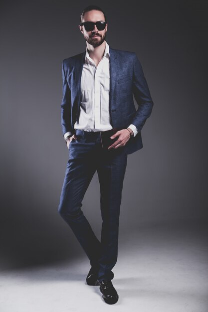 Retrato de moda guapo elegante hipster empresario modelo vestido con elegante traje azul con gafas de sol posando en gris