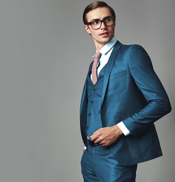 Retrato de moda guapo elegante hipster empresario modelo vestido con elegante traje azul posando sobre fondo gris en estudio en gafas