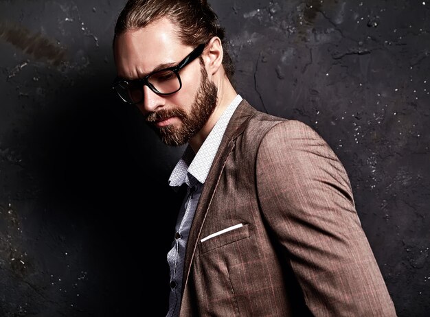 Retrato de moda guapo elegante hipster empresario modelo vestido con elegante traje marrón con gafas cerca de la pared oscura