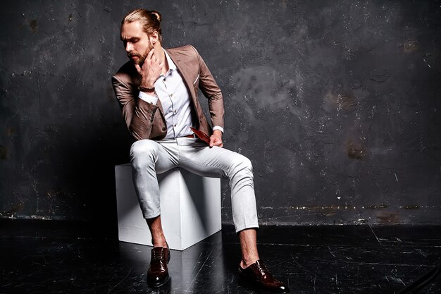 Retrato de moda guapo elegante hipster empresario modelo vestido con elegante traje marrón sentado cerca de la oscuridad en el estudio