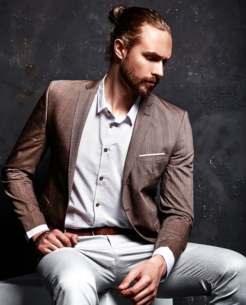 Retrato de moda guapo elegante hipster empresario modelo vestido con elegante traje marrón sentado cerca de la oscuridad