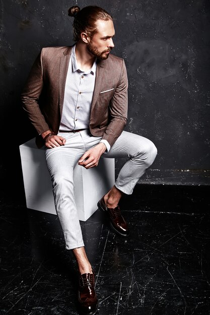 Retrato de moda guapo elegante hipster empresario modelo vestido con elegante traje marrón sentado cerca de la oscuridad