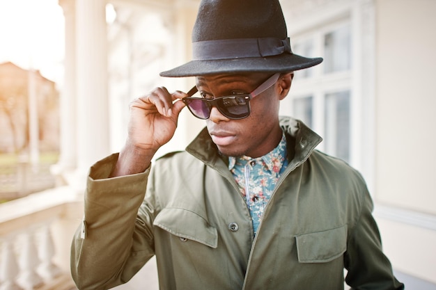 Retrato de moda de hombre afroamericano negro con capa de abrigo verde y sombrero negro en el balcón de la mansión amarilla