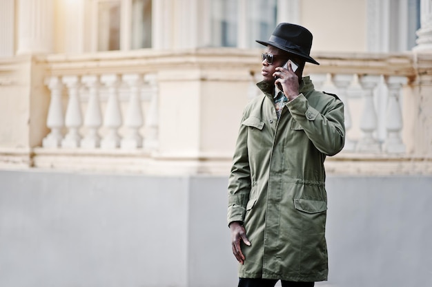 Foto gratuita retrato de moda de un hombre afroamericano negro con capa de abrigo verde y sombrero negro hablando en la mansión amarilla de fondo del teléfono móvil