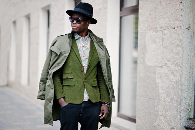 Retrato de moda de hombre afroamericano negro con chaqueta de terciopelo verde, sombrero negro y capa de abrigo sobre los hombros caminando por las calles de la casa de fondo de la ciudad con muchas ventanas