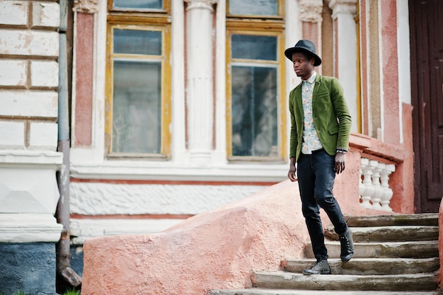 Retrato de moda de un hombre afroamericano negro con una chaqueta de terciopelo verde y un sombrero negro que se queda en las escaleras en la vieja mansión de fondo