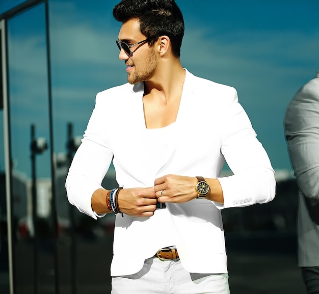 Retrato de moda de hombre joven guapo modelo sexy en traje blanco casual en gafas de sol en traje blanco en la calle